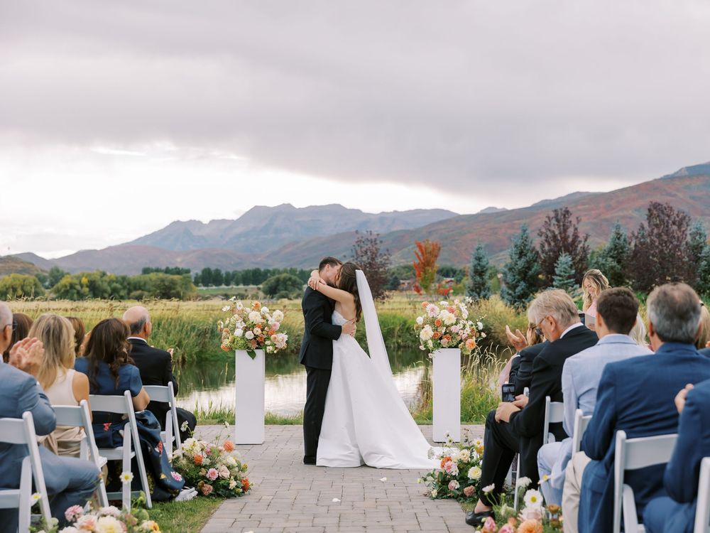 Bride and groom share first kiss at outdoor wedding ceremony with mountain backdrop and floral arrangements.