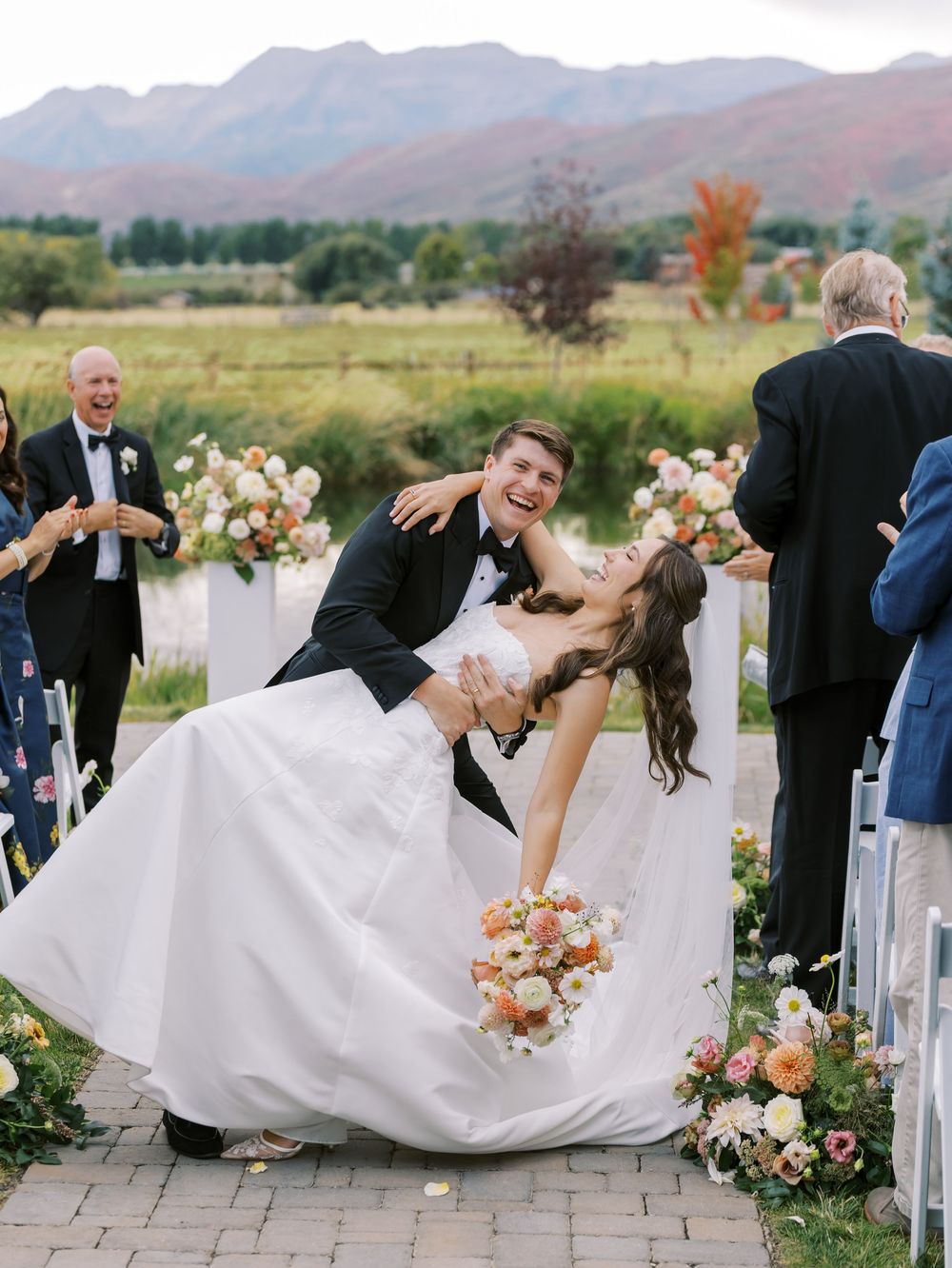 Joyful newlyweds share a dip kiss after outdoor wedding ceremony with mountain backdrop and colorful floral arrangements.