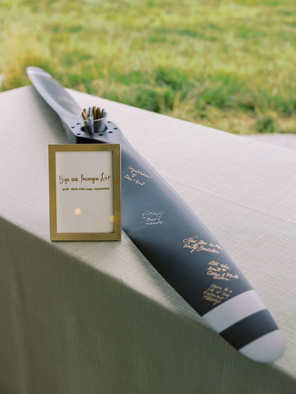A dark airplane propeller used as a unique wedding guest book, signed with gold ink, displayed on a draped table outdoors.