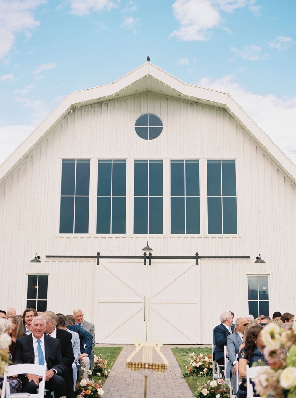 Outdoor wedding ceremony in front of a white barn venue with large windows, floral arrangements, and seated guests on a sunny day.