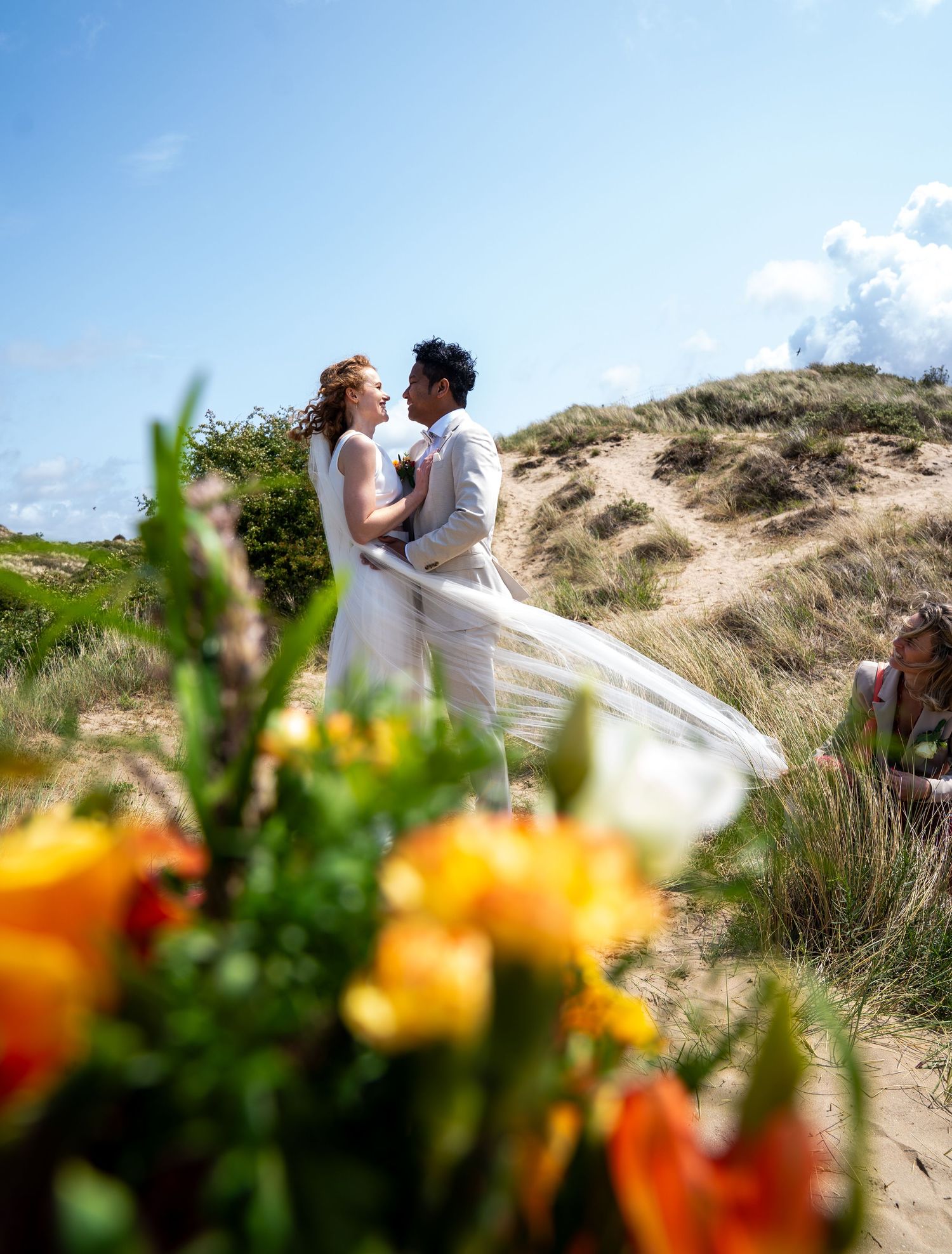 Een bruidspaar omhelst elkaar in de zandduinen met kleurrijke wilde bloemen op de voorgrond.