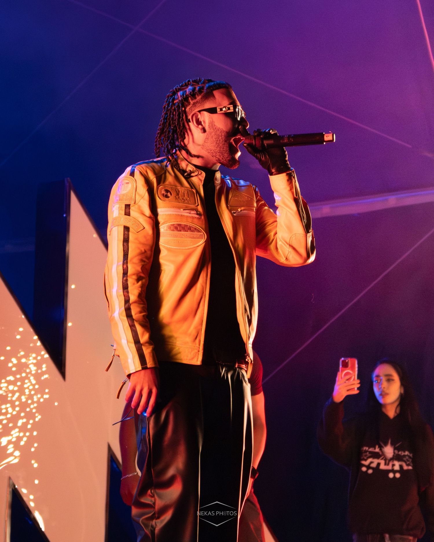 A rapper performs on stage holding a microphone, wearing a tan jacket, with purple stage lighting and sparks in the background.
