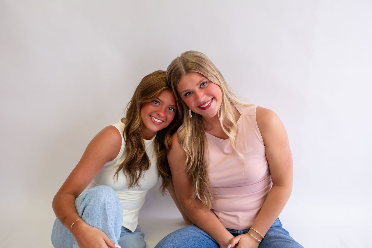 Two smiling young women with long hair sit together against a white background, wearing casual tops and jeans.