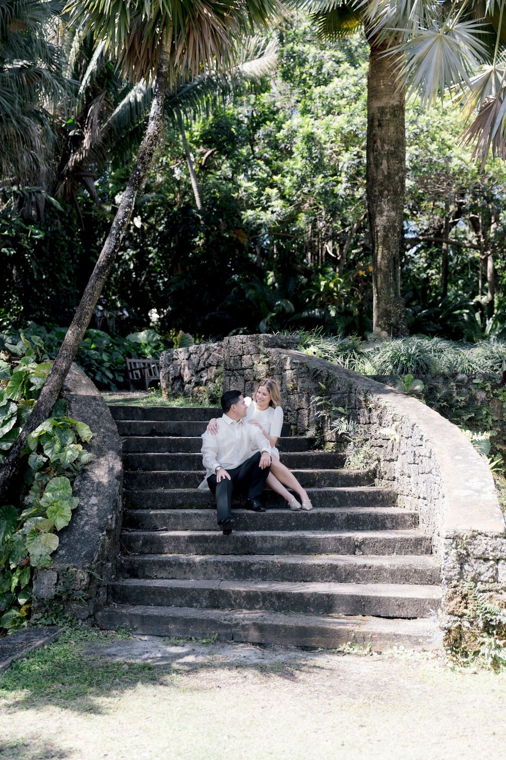 Couple sitting closely together on stone steps surrounded by lush tropical greenery and palm trees in a garden setting.