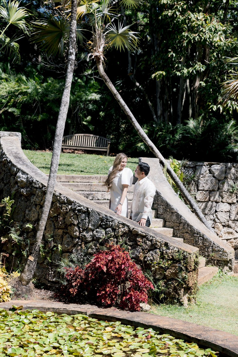 Couple holding hands on stone steps beside a lily pond in a lush tropical garden.