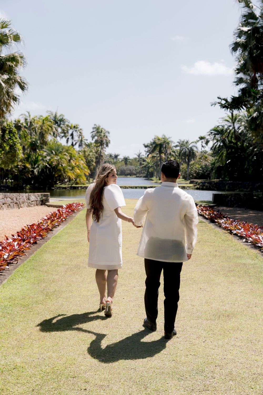Couple in white outfits holding hands while walking along a lush garden path lined with tropical trees and colorful plants.