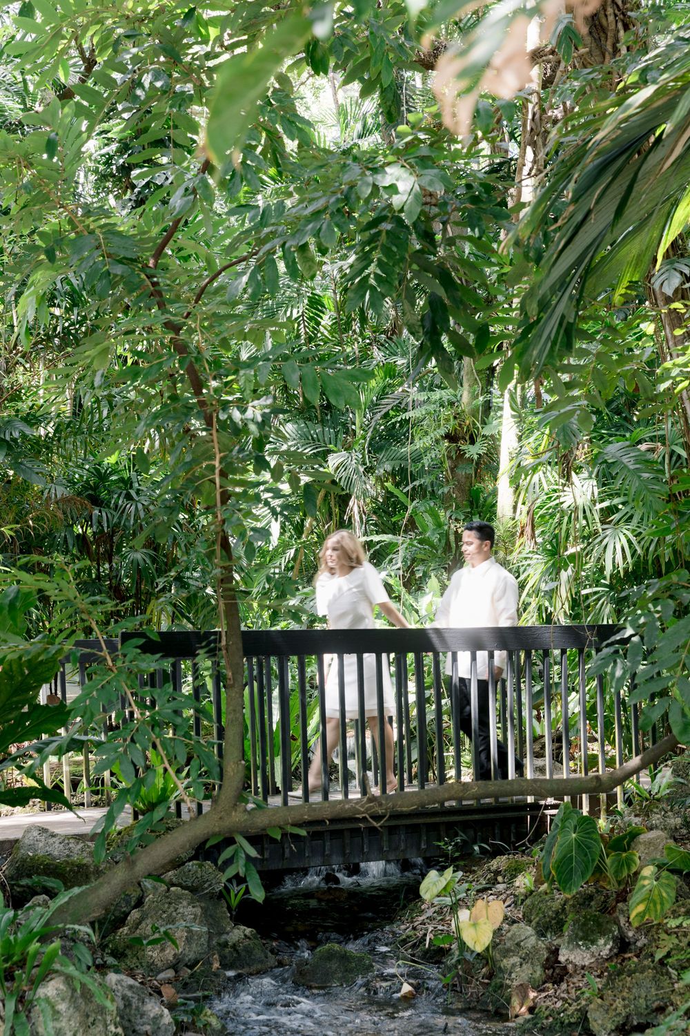 Couple in white attire standing on a wooden bridge surrounded by lush tropical greenery.