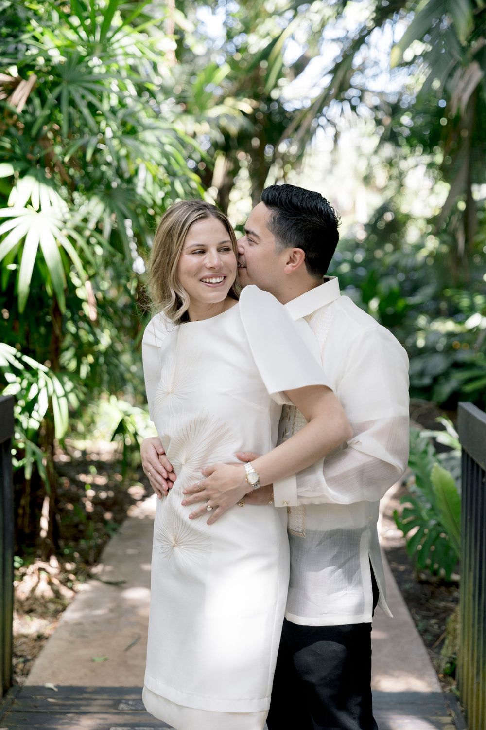 Smiling couple embracing on a garden path, man kissing woman's cheek in tropical setting.