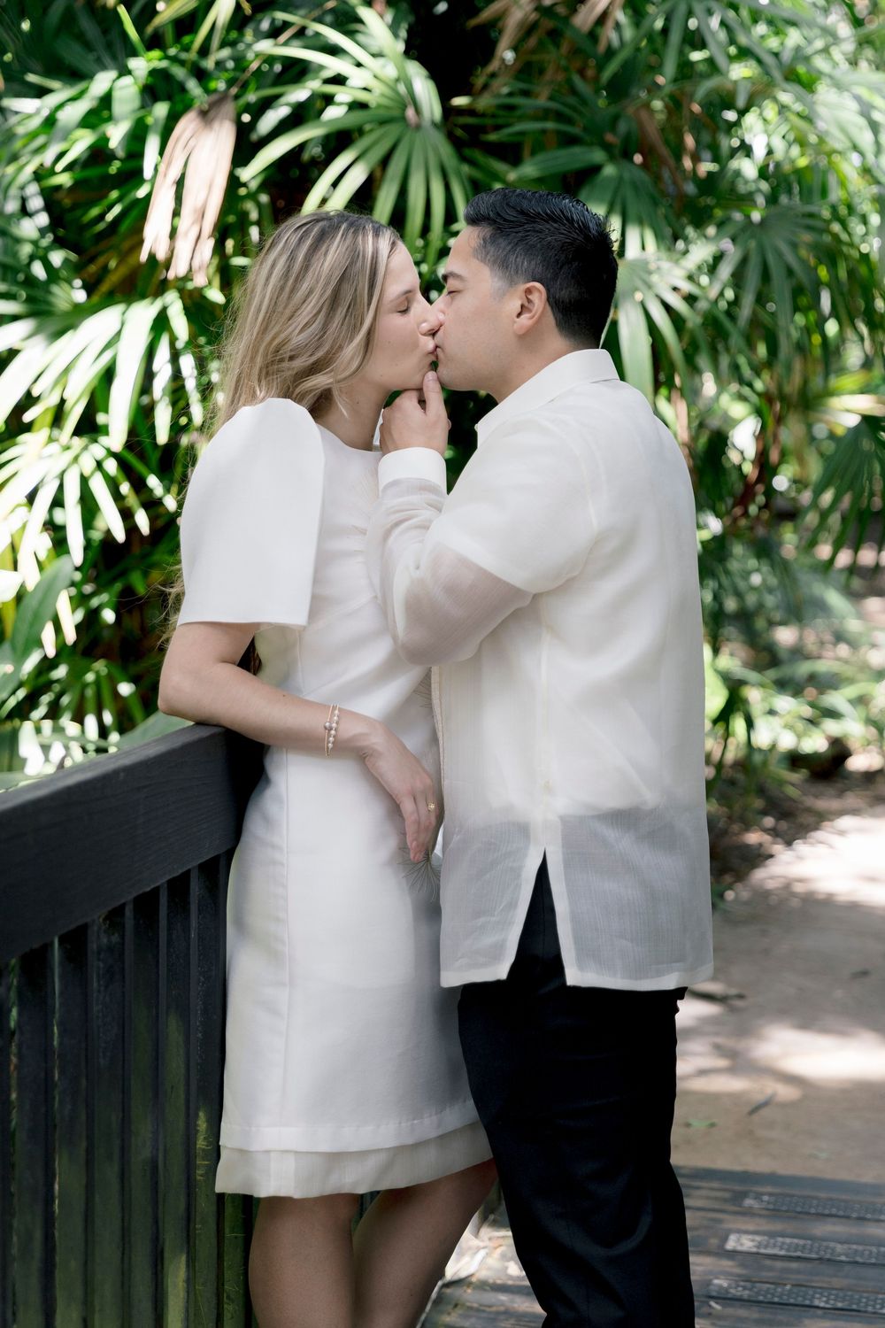 Couple sharing a romantic kiss in a lush tropical garden, woman in white dress, man in barong tagalog.
