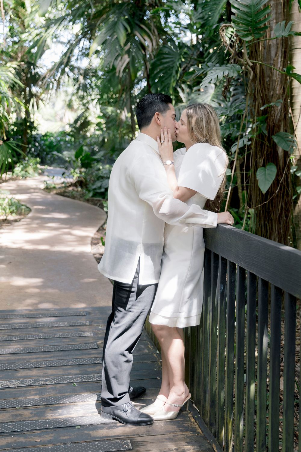 Couple in white outfits sharing a kiss on a wooden bridge surrounded by lush tropical greenery.