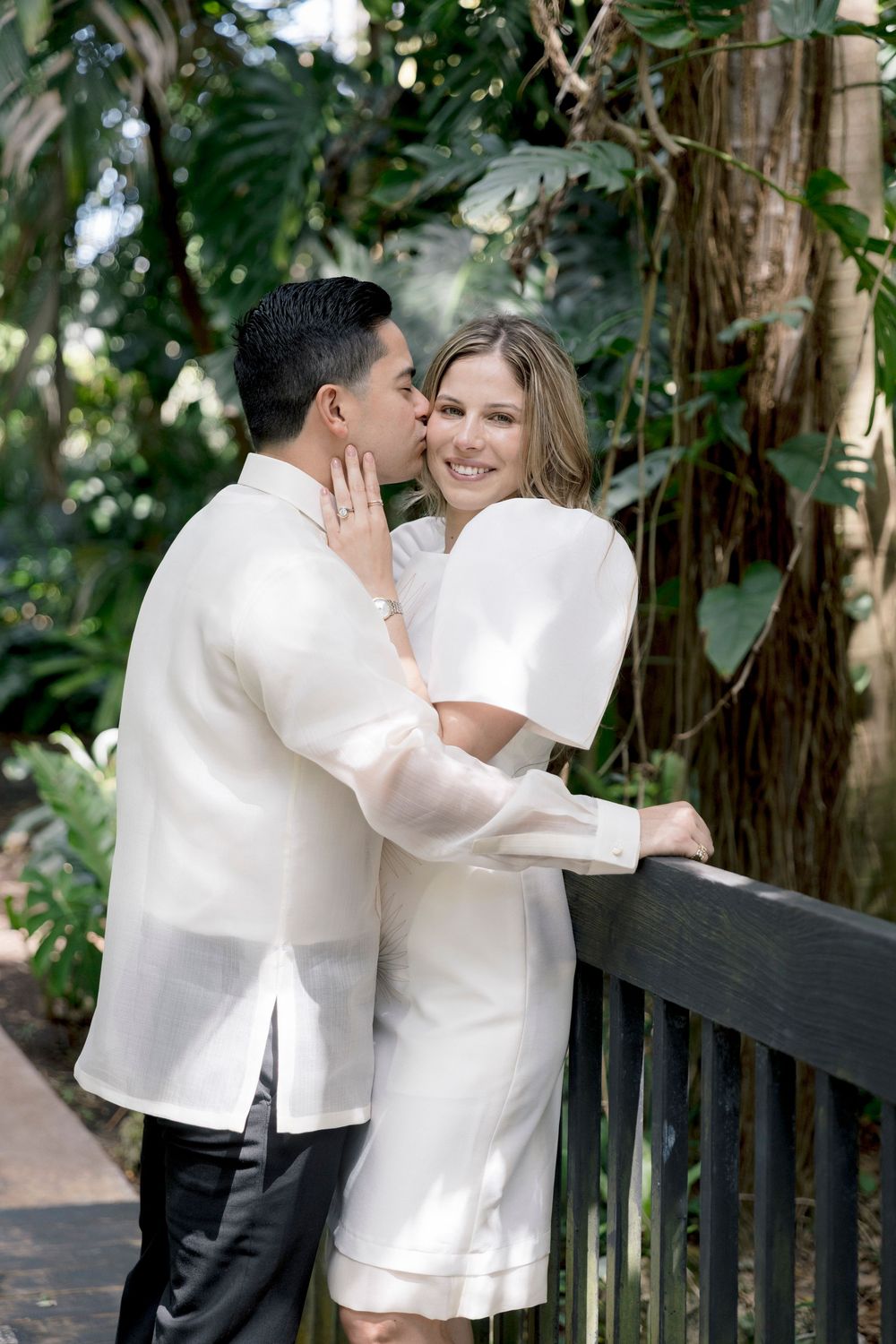 Couple in white wedding attire embracing on a wooden bridge surrounded by lush tropical greenery.