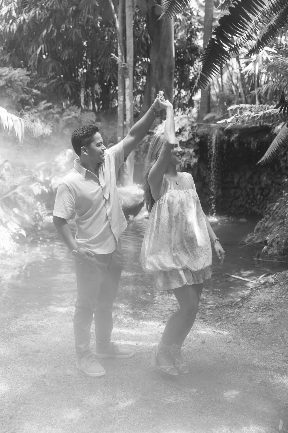 A couple dances joyfully in a lush tropical garden, the man spinning the woman in a sundress, captured in black and white.