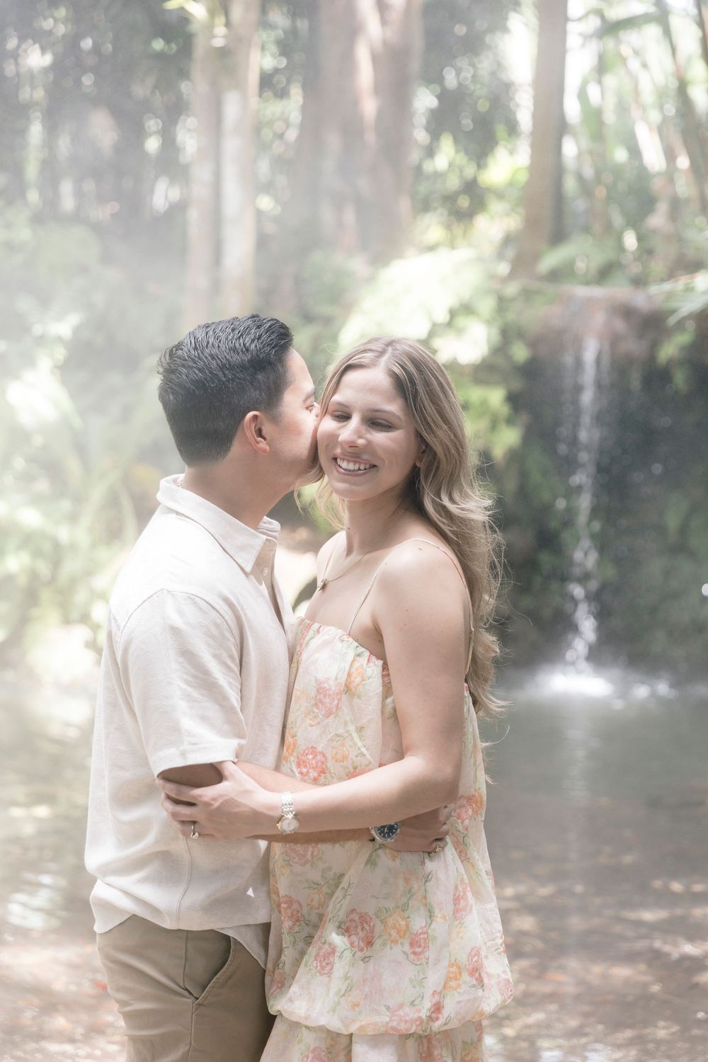 Couple embracing near a misty waterfall in a serene forest setting.