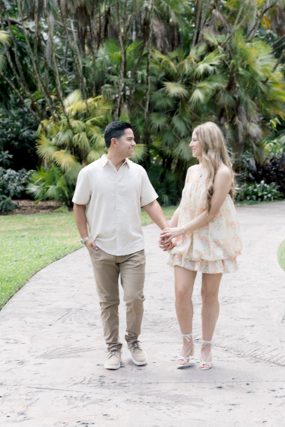 A couple holds hands and smiles at each other while walking along a path surrounded by tropical greenery.