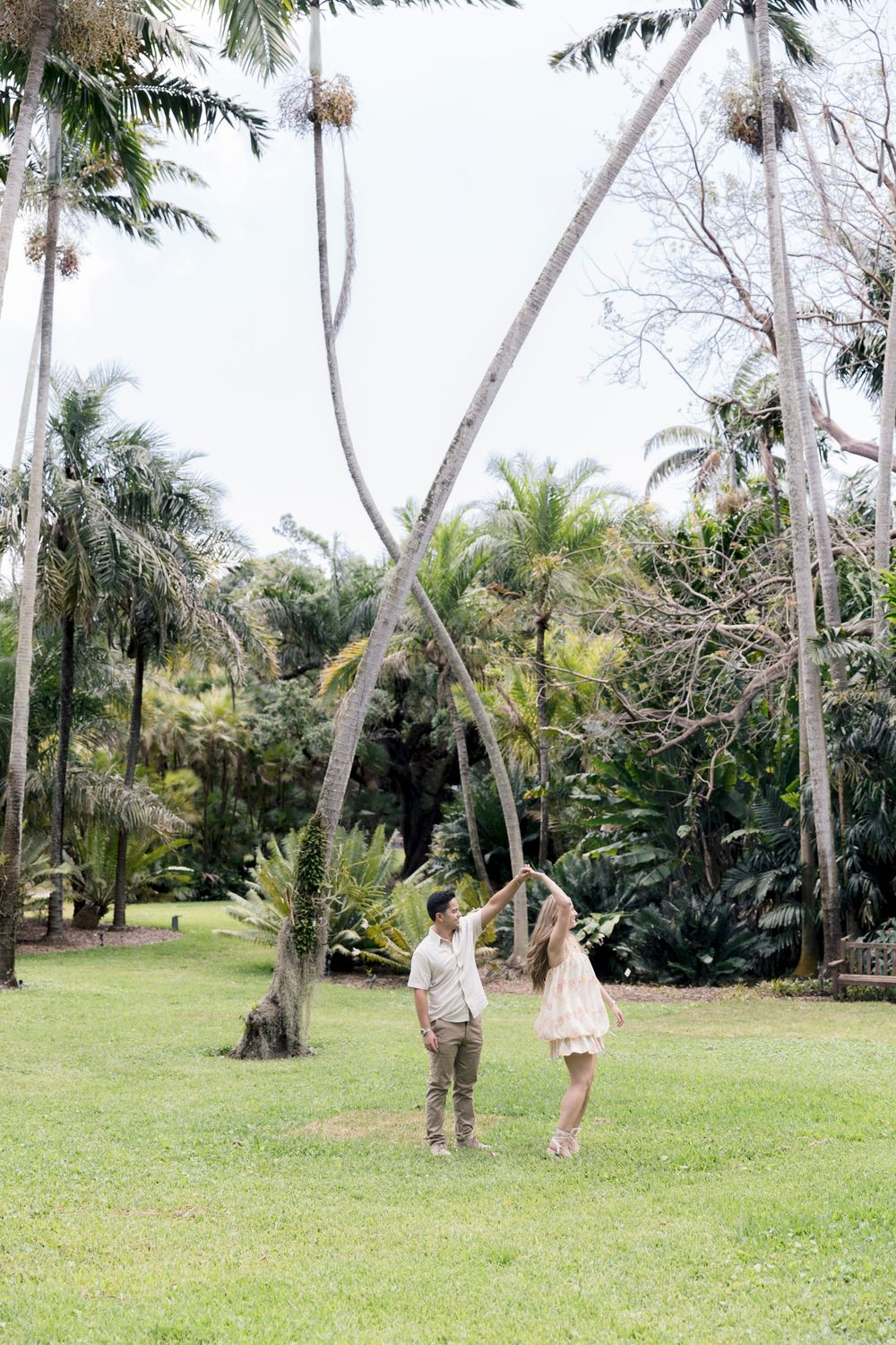 Couple dancing together on green lawn surrounded by tall tropical palm trees in a lush garden setting.