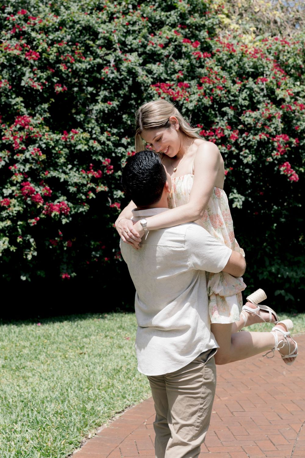 Man lifting woman in a joyful embrace on a brick path with red flowers behind them.