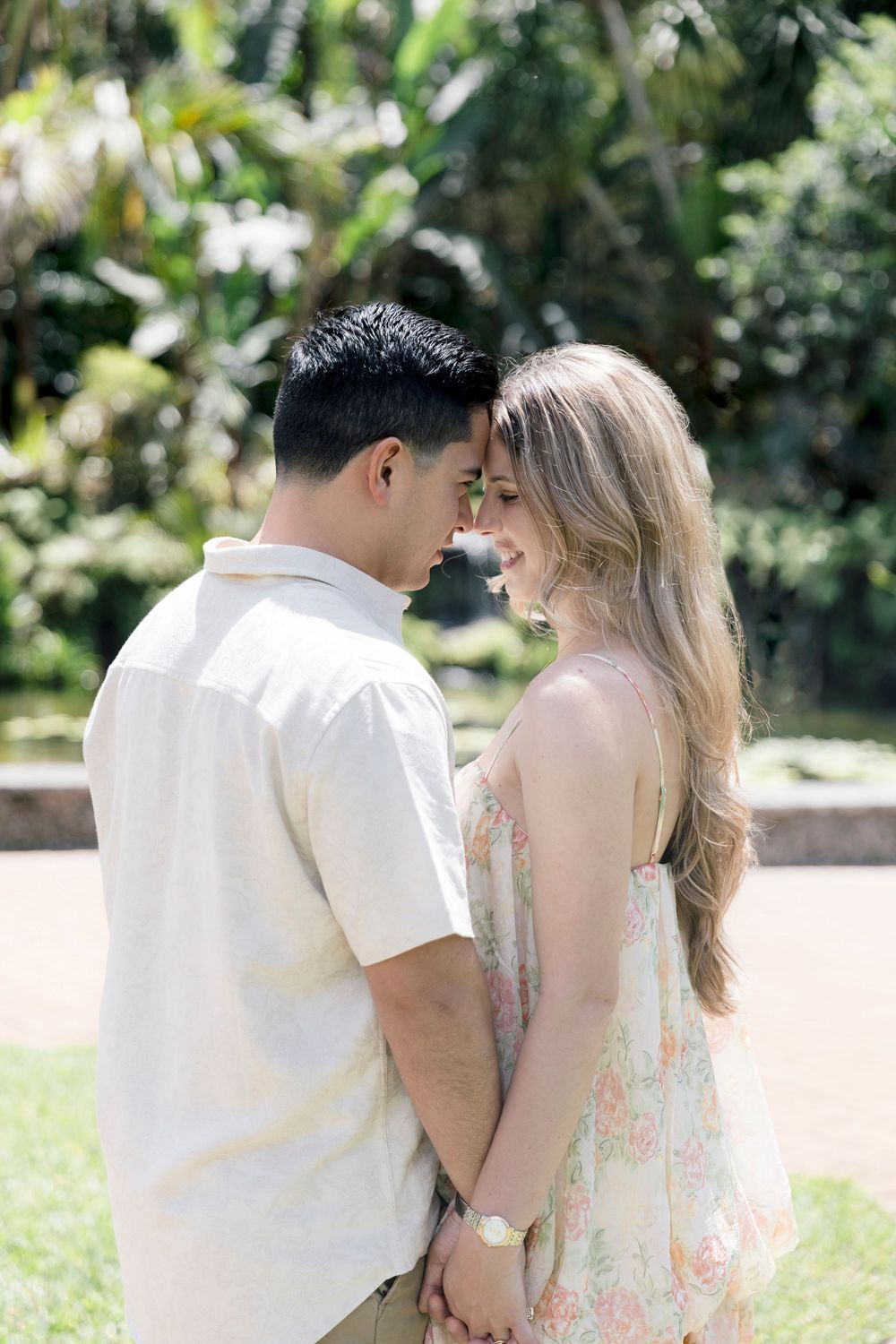 A couple holds hands and touches foreheads while smiling in a lush tropical garden setting.