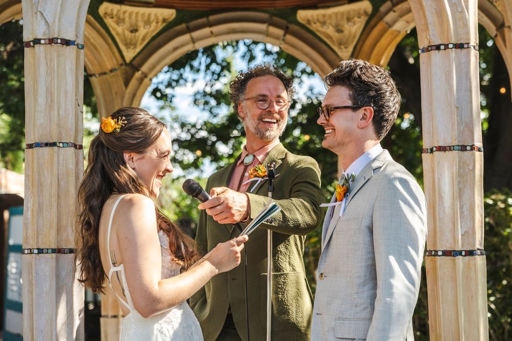 Couple laughing with officiant during outdoor wedding ceremony under wooden arbor with lush green trees.