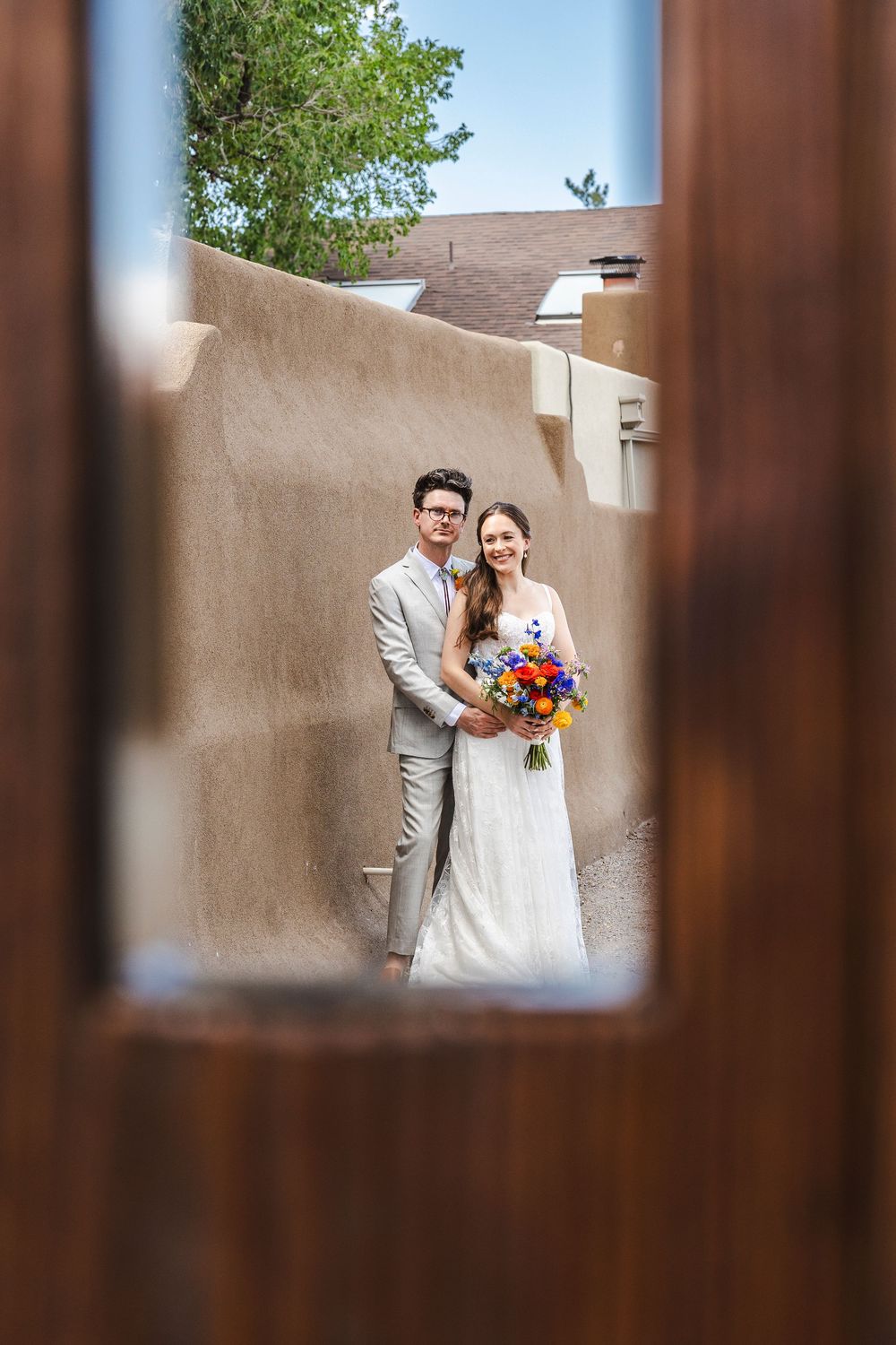 Bride and groom pose together framed by wooden doors, adobe walls backdrop, colorful bouquet.
