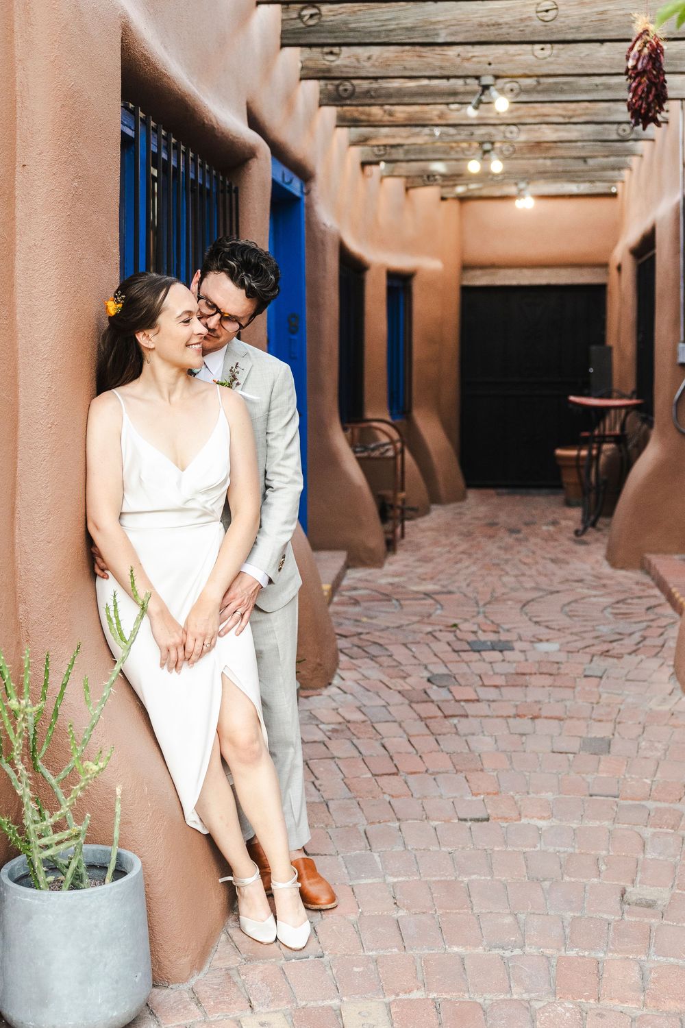Couple in wedding attire embracing in a Southwest adobe courtyard with brick walkway and string lights.
