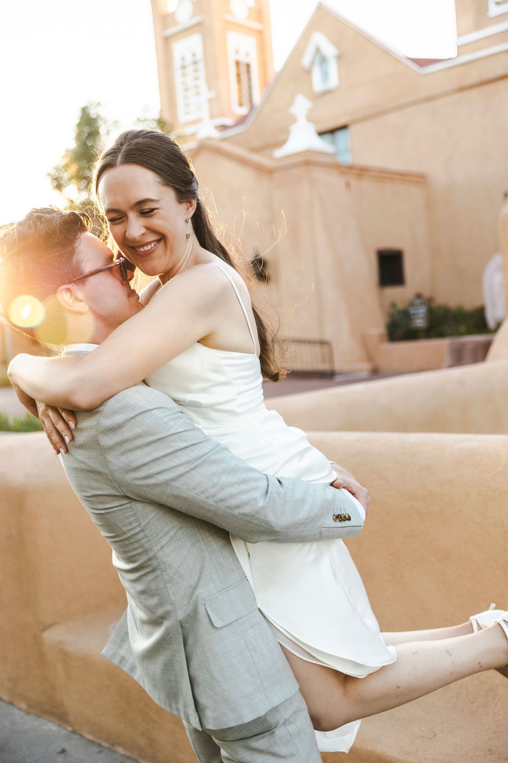 Joyful bride in white dress being lifted by groom in gray suit outside adobe building at golden hour.