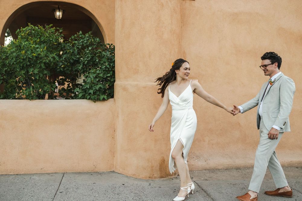 Bride and groom walk hand-in-hand laughing outside adobe building on sunny wedding day.