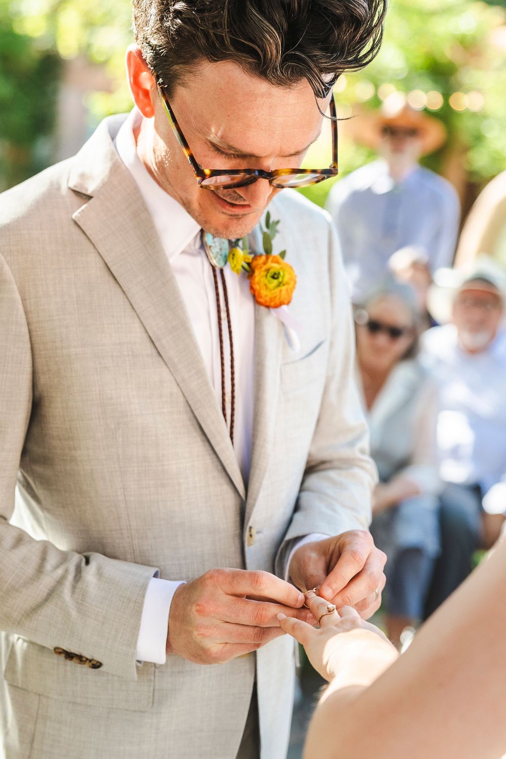 Groom in beige suit with orange boutonniere placing wedding ring on bride's finger during outdoor ceremony.