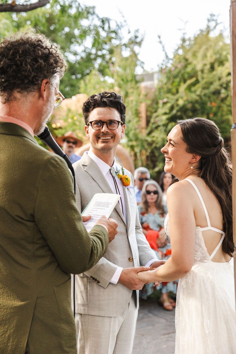 A joyful outdoor wedding ceremony with bride and groom holding hands, officiant smiling, guests seated in background.