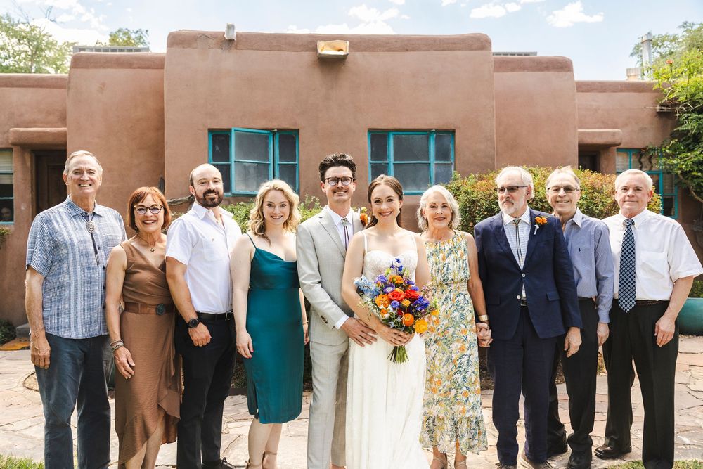 Wedding party poses in front of adobe building, bride holding colorful bouquet surrounded by smiling family members outdoors.