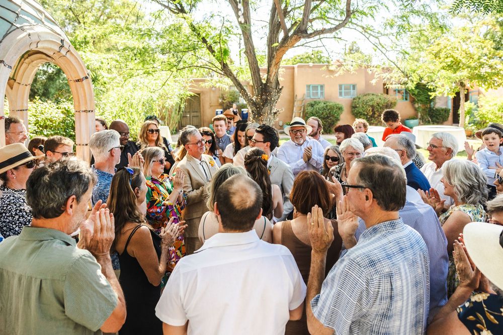 Large outdoor gathering of guests mingling and applauding at a sunny garden event near adobe architecture.