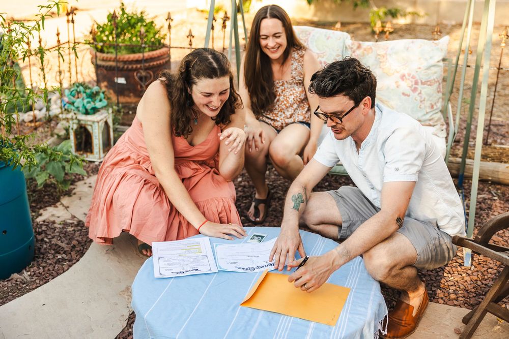 Three friends laugh together reviewing documents at an outdoor patio table in a sunny garden setting.