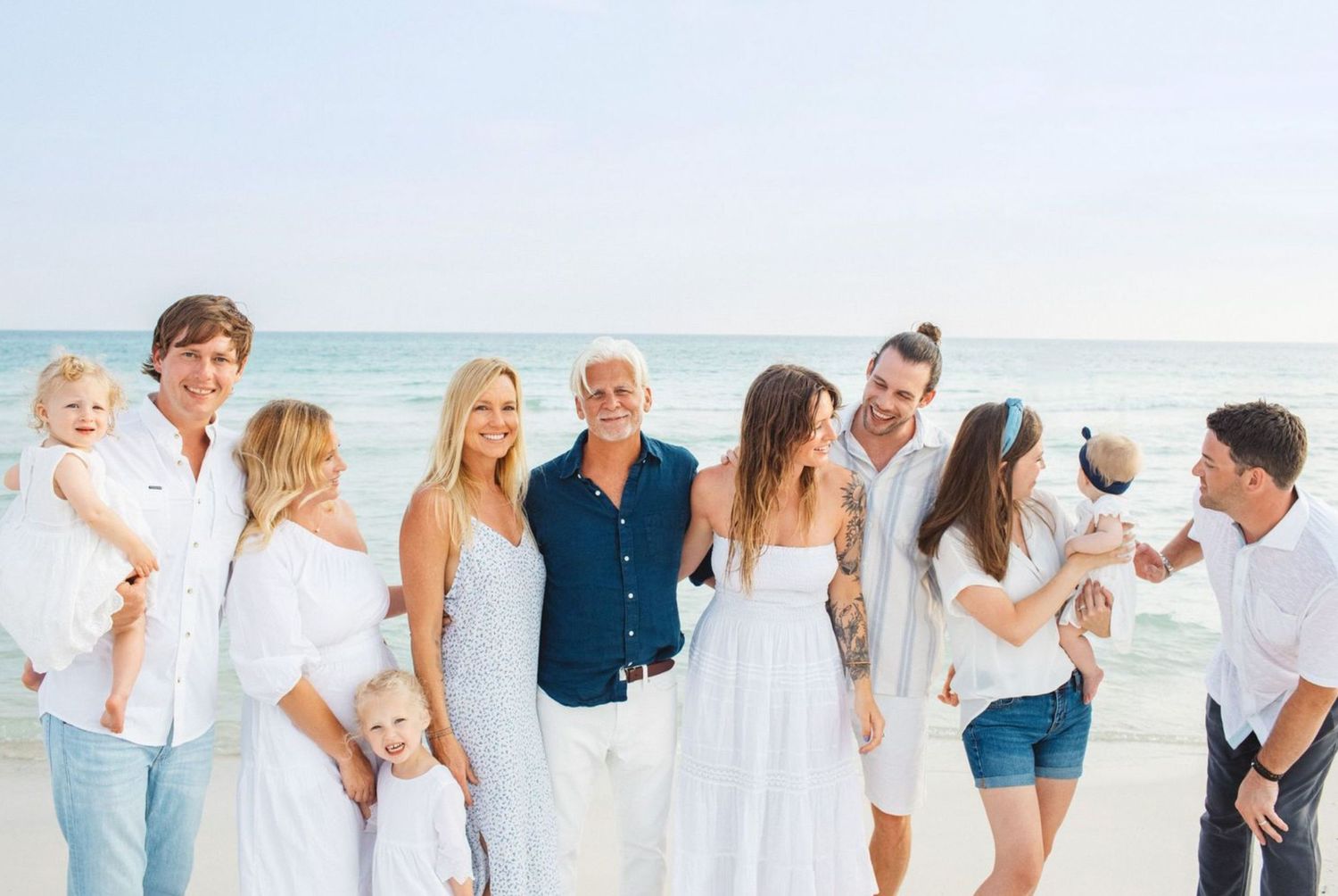 Large family group photo on a sunny beach, wearing coordinated white outfits, smiling together near the ocean.