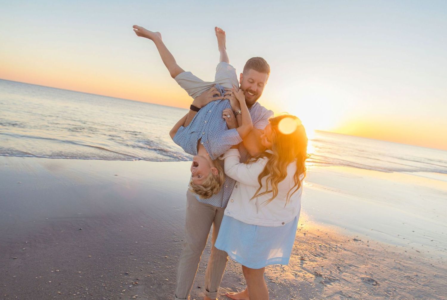Family playing on beach at sunset, father lifting child while mother watches, golden hour light.