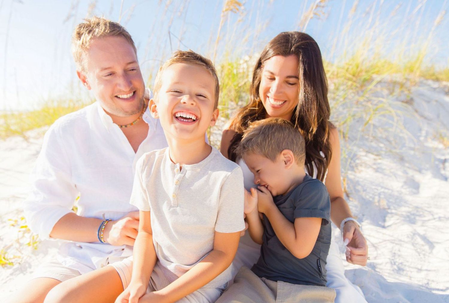 Happy family of four laughing together on a sunny beach with white sand dunes in the background.