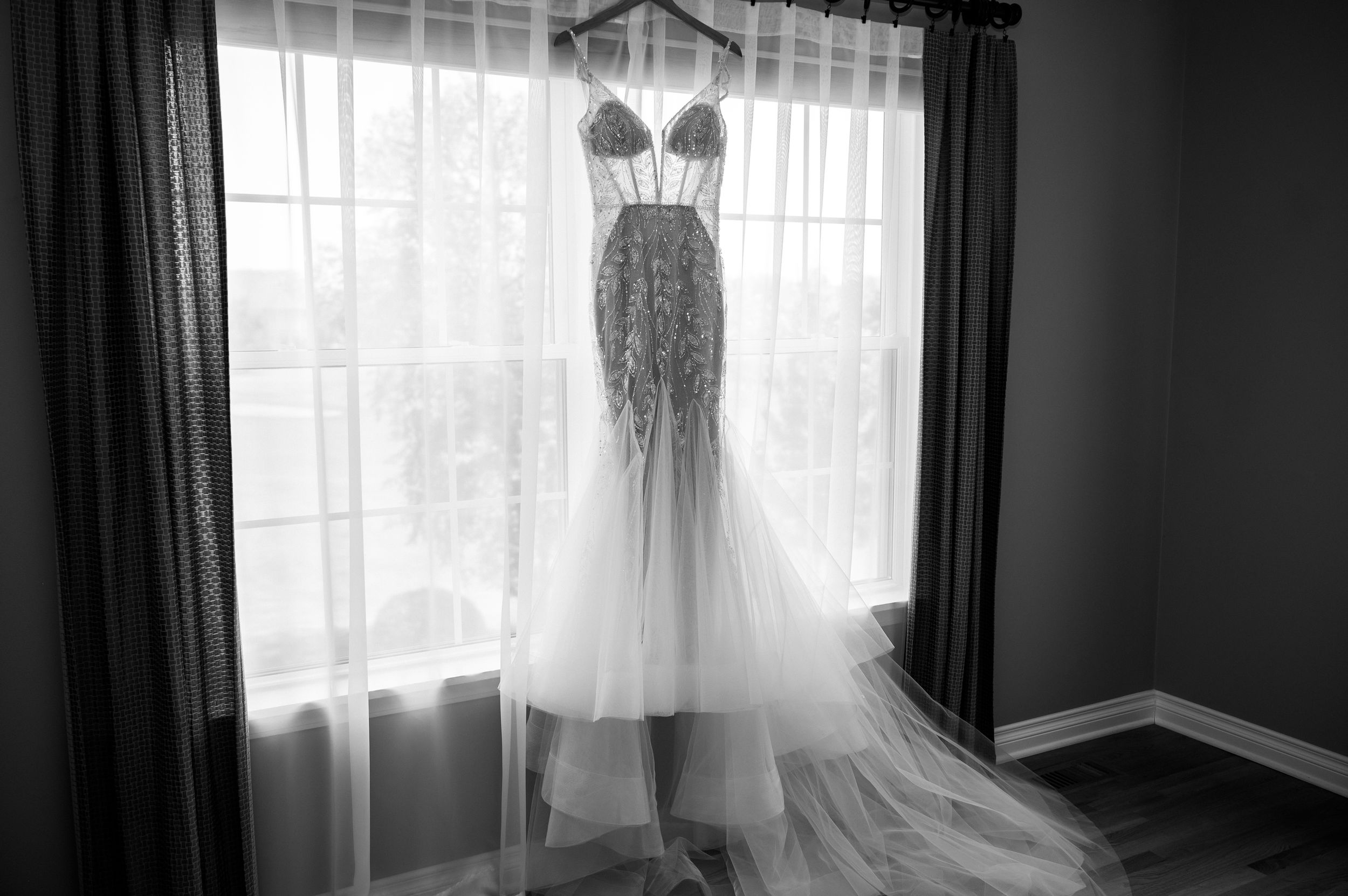 Elegant beaded mermaid wedding dress hanging in window, black and white bridal gown photography.
