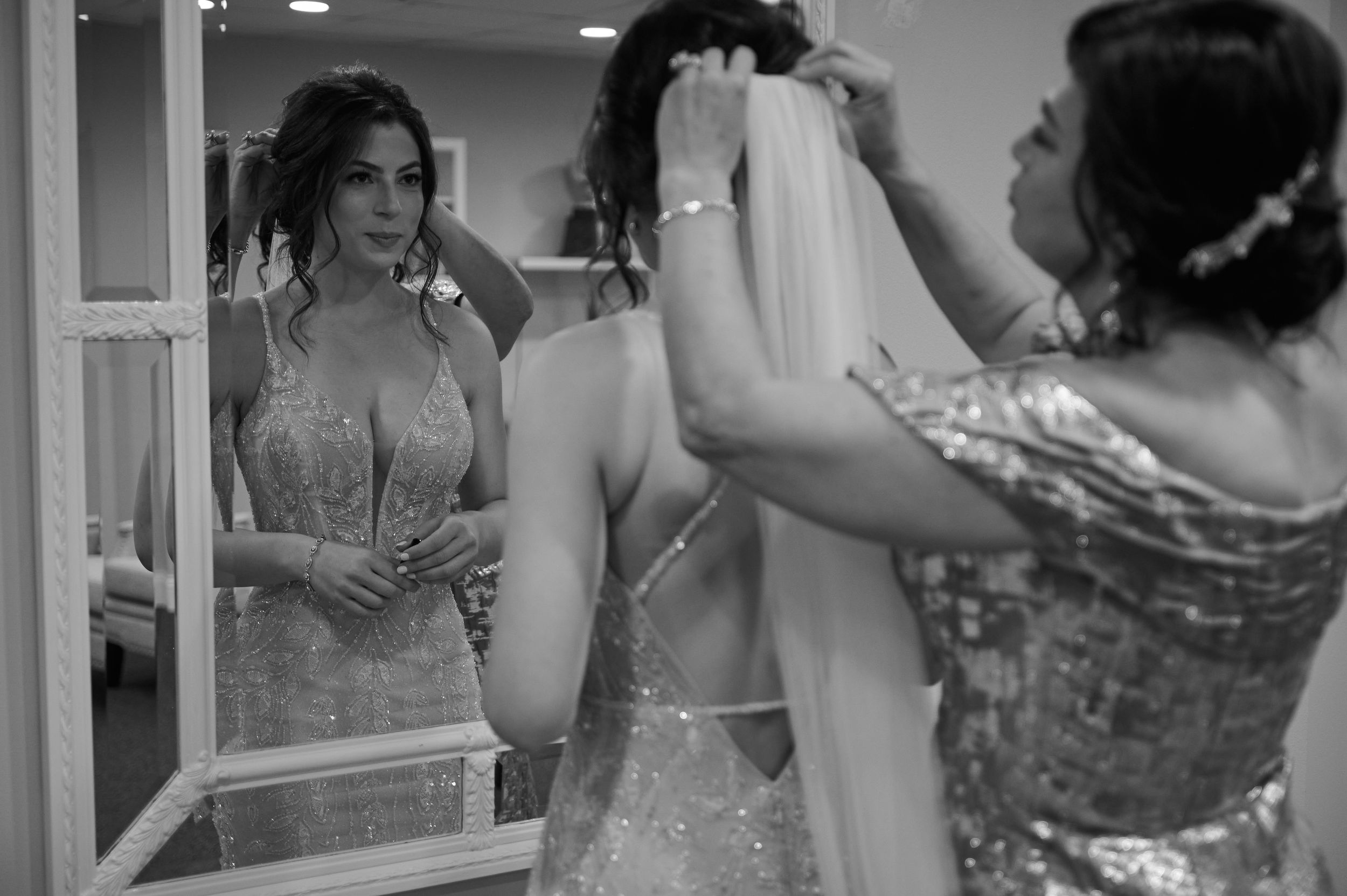 Black and white photo of bride in beaded gown getting veil adjusted, reflected in mirror by bridesmaid.