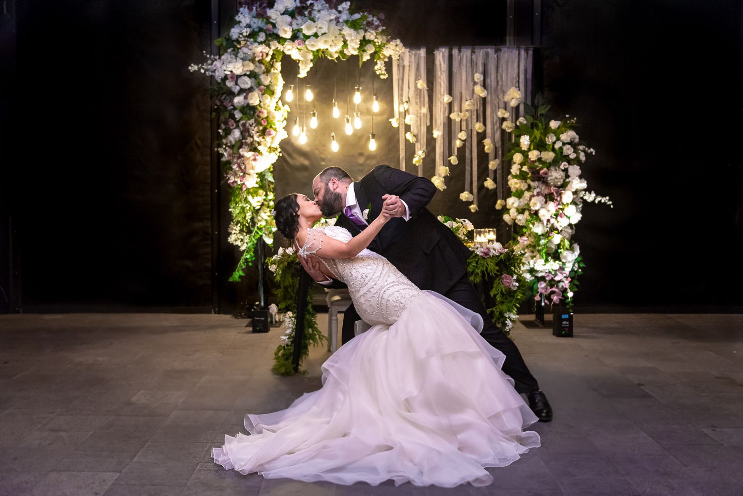 Couple shares passionate dip kiss in front of white floral arch with warm glowing lights.