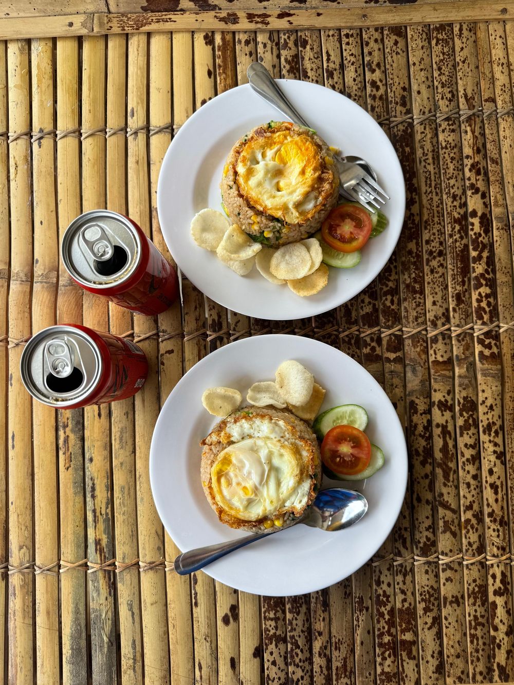 Two plates of Indonesian nasi goreng fried rice topped with fried eggs, served with fresh vegetables and canned drinks.