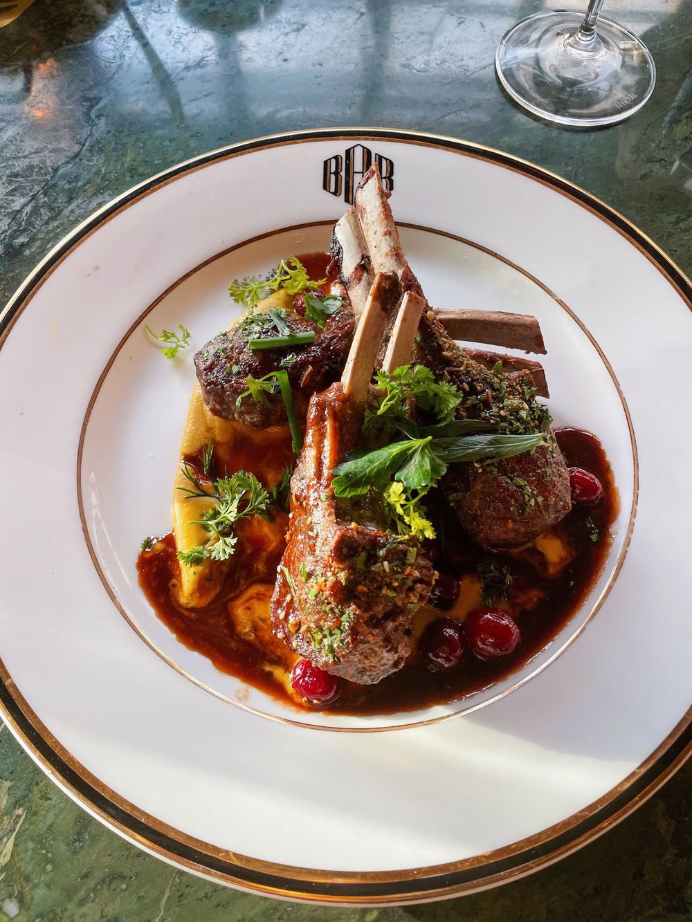 Elegant plated lamb chops with herb crust, red wine sauce, and garnishes on a gold-rimmed monogrammed white plate.