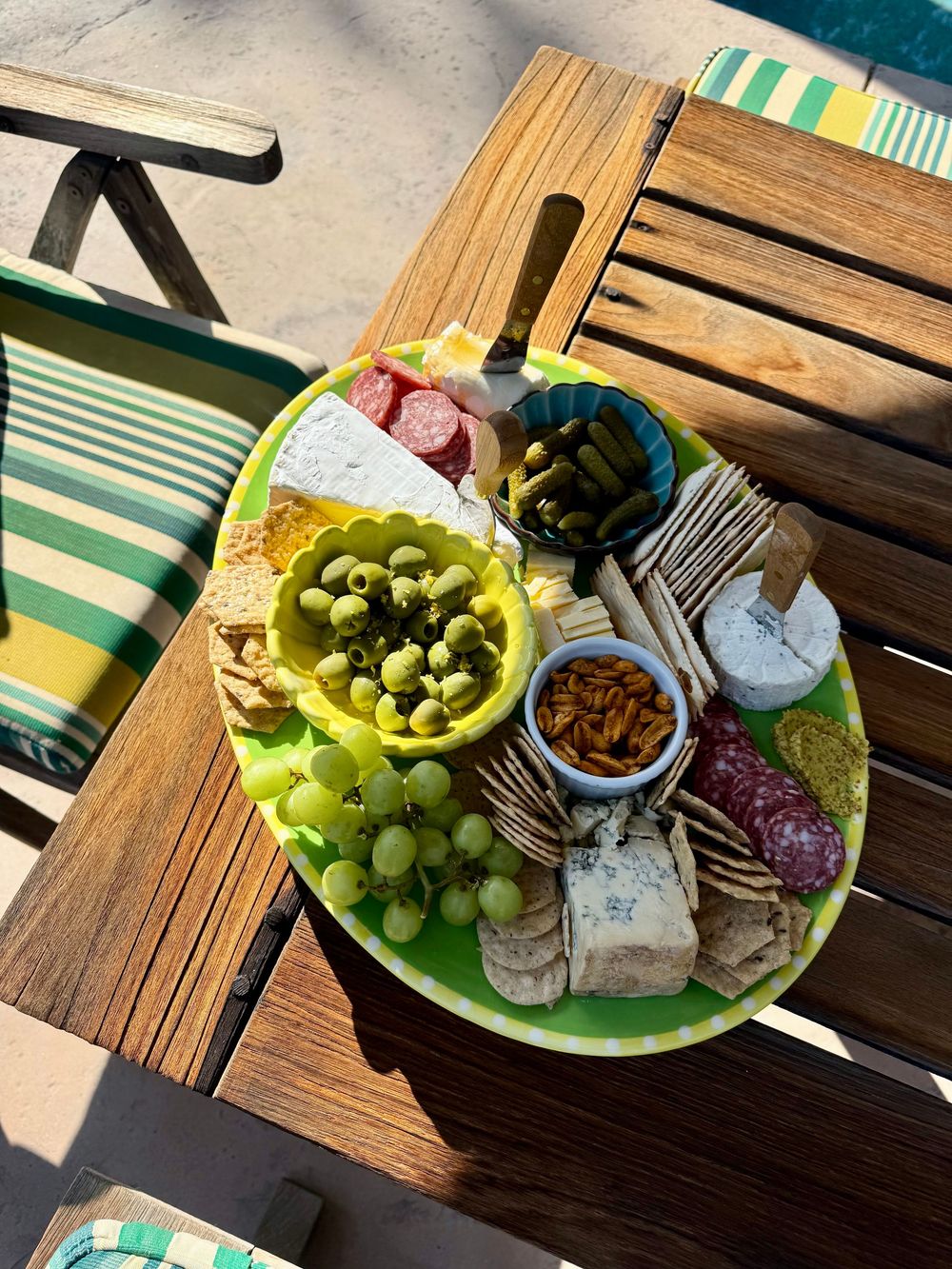 Overhead view of a colorful charcuterie platter with grapes, cheese, salami, crackers, pickles, and olives on a wooden table.