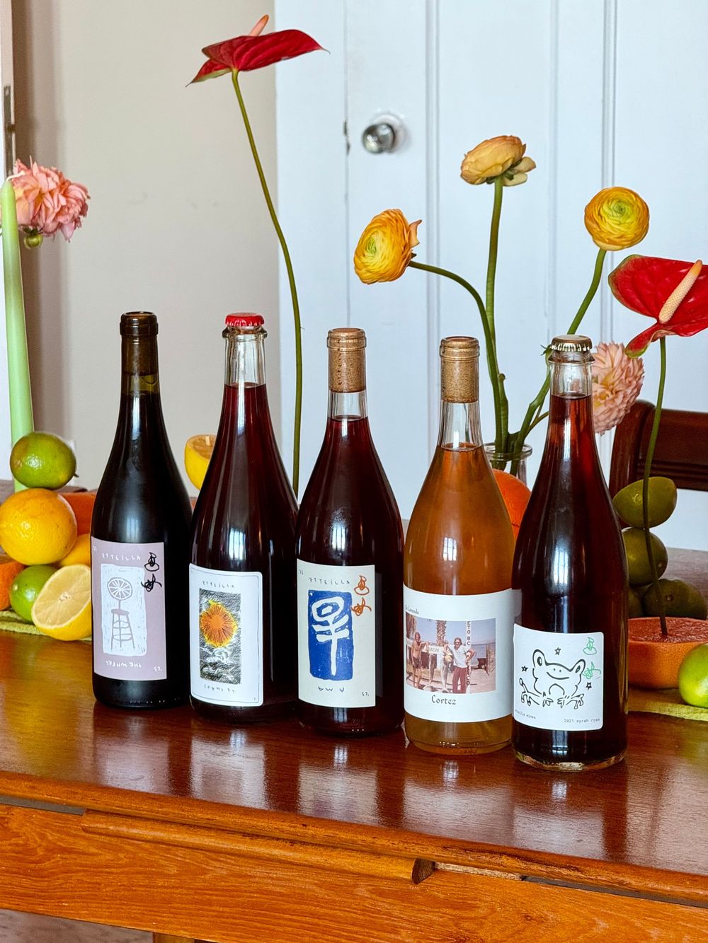 Five natural wine bottles with artistic labels displayed on a wooden table with fresh flowers and citrus fruit.