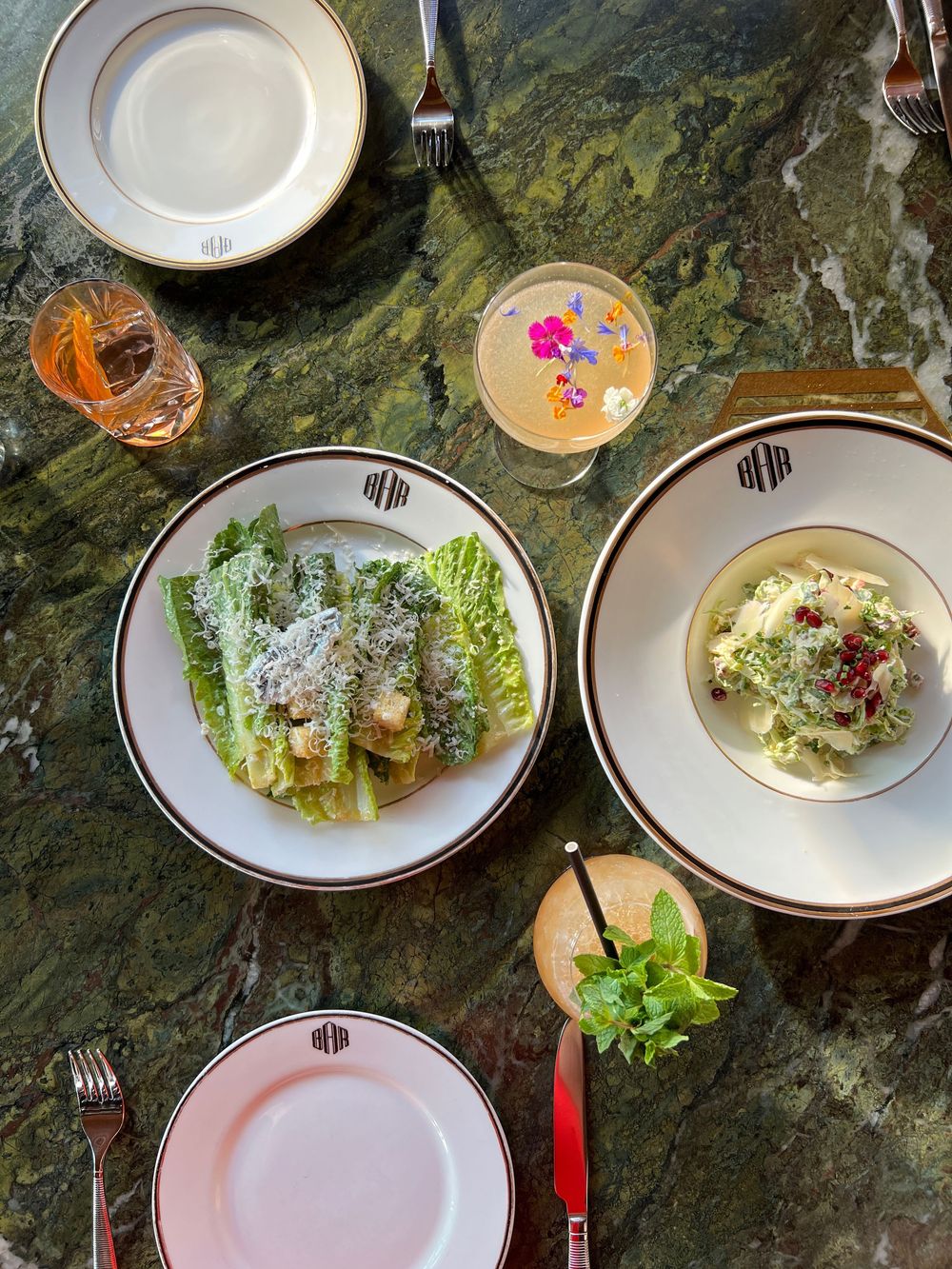 Elegant restaurant table with Caesar salad, pasta dish, cocktails, and monogrammed plates on green marble.