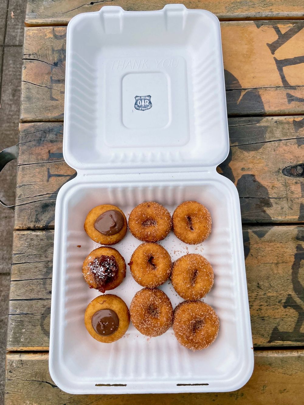 Nine mini donuts in a styrofoam takeout container, some coated in cinnamon sugar and others topped with chocolate glaze.