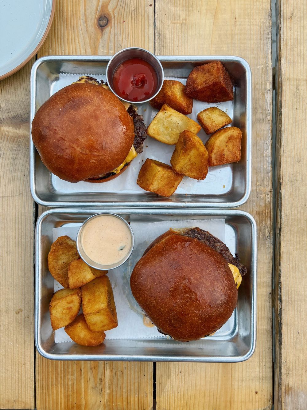 Two metal trays with juicy burgers on brioche buns, golden roasted potato cubes, and dipping sauces on a wooden table.