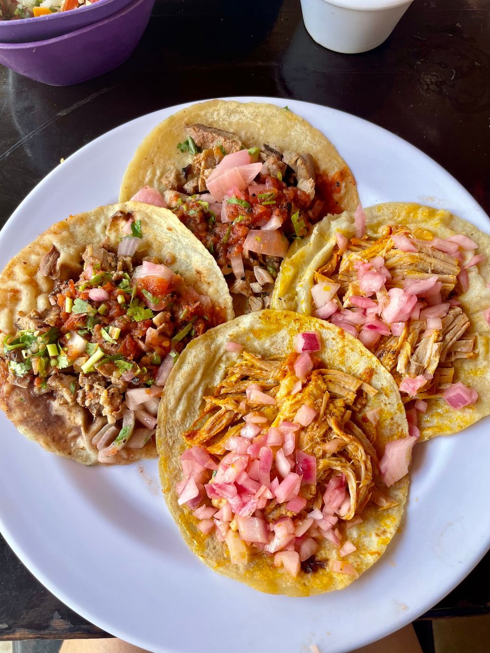 Four authentic Mexican street tacos on a white plate topped with pickled onions, cilantro, and salsa.