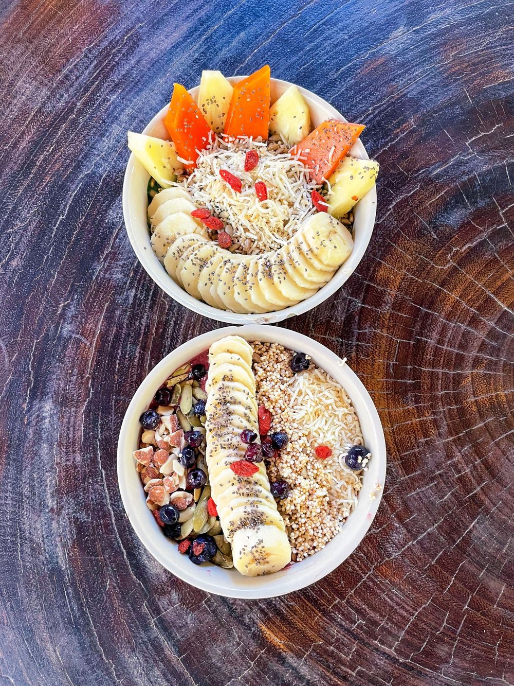 Two healthy acai bowls topped with banana, granola, goji berries, blueberries, and fresh fruit on a wood surface.