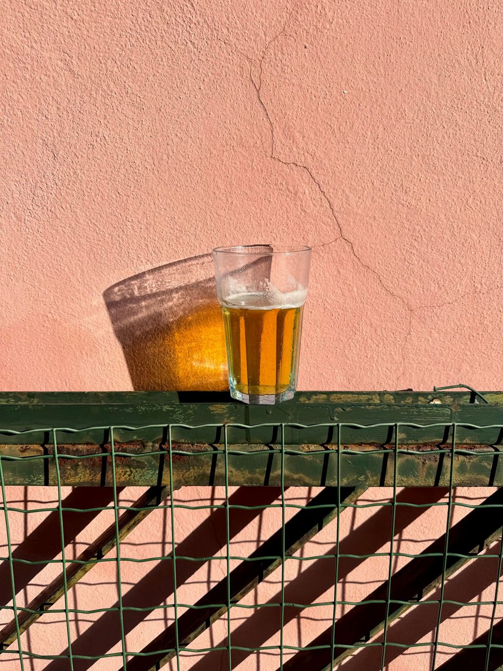Glass of golden beer casting a shadow on a green metal railing against a textured pink stucco wall.
