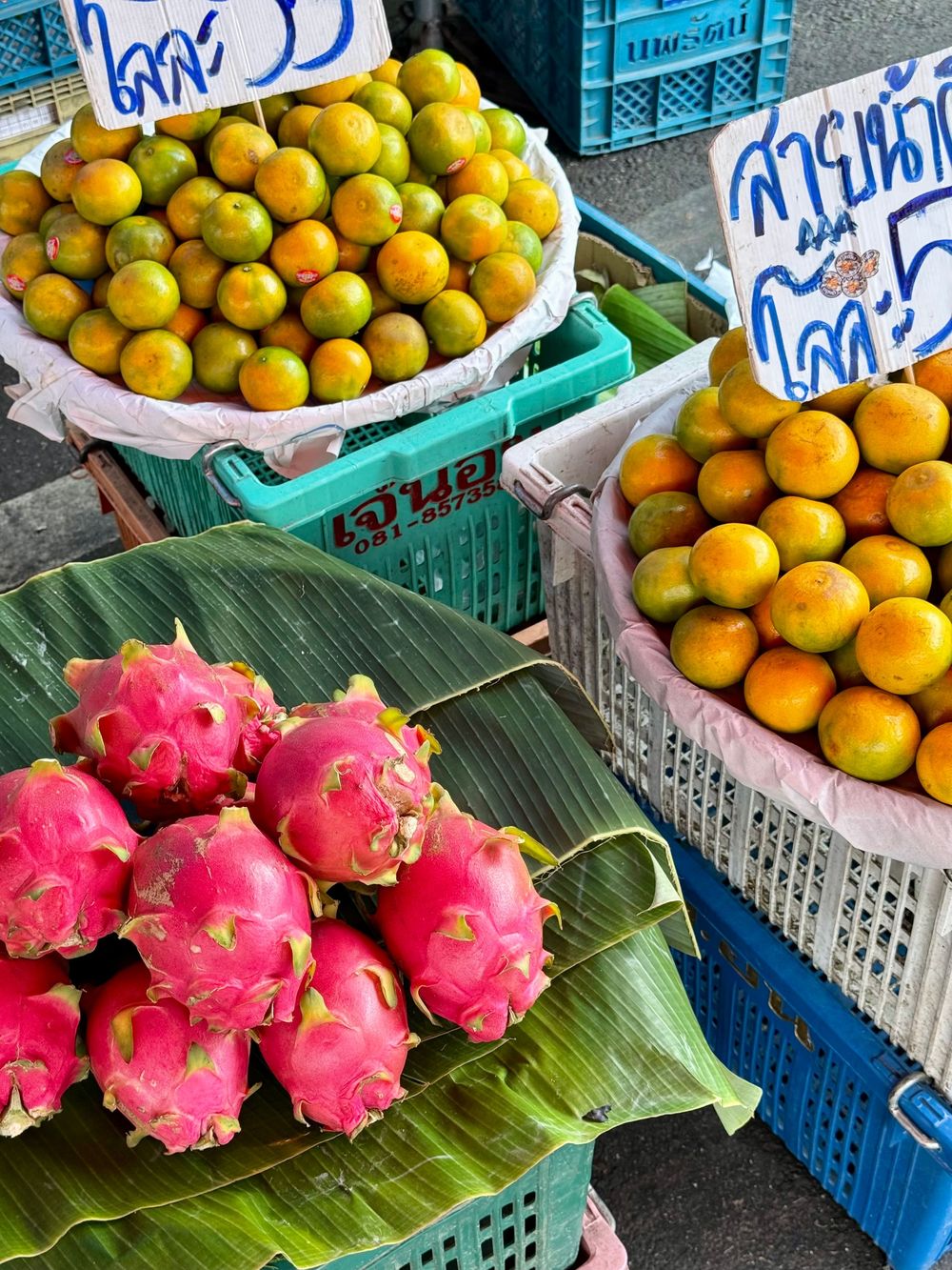 Colorful Thai market display of fresh dragon fruit on banana leaves and orange mandarins in blue crates.