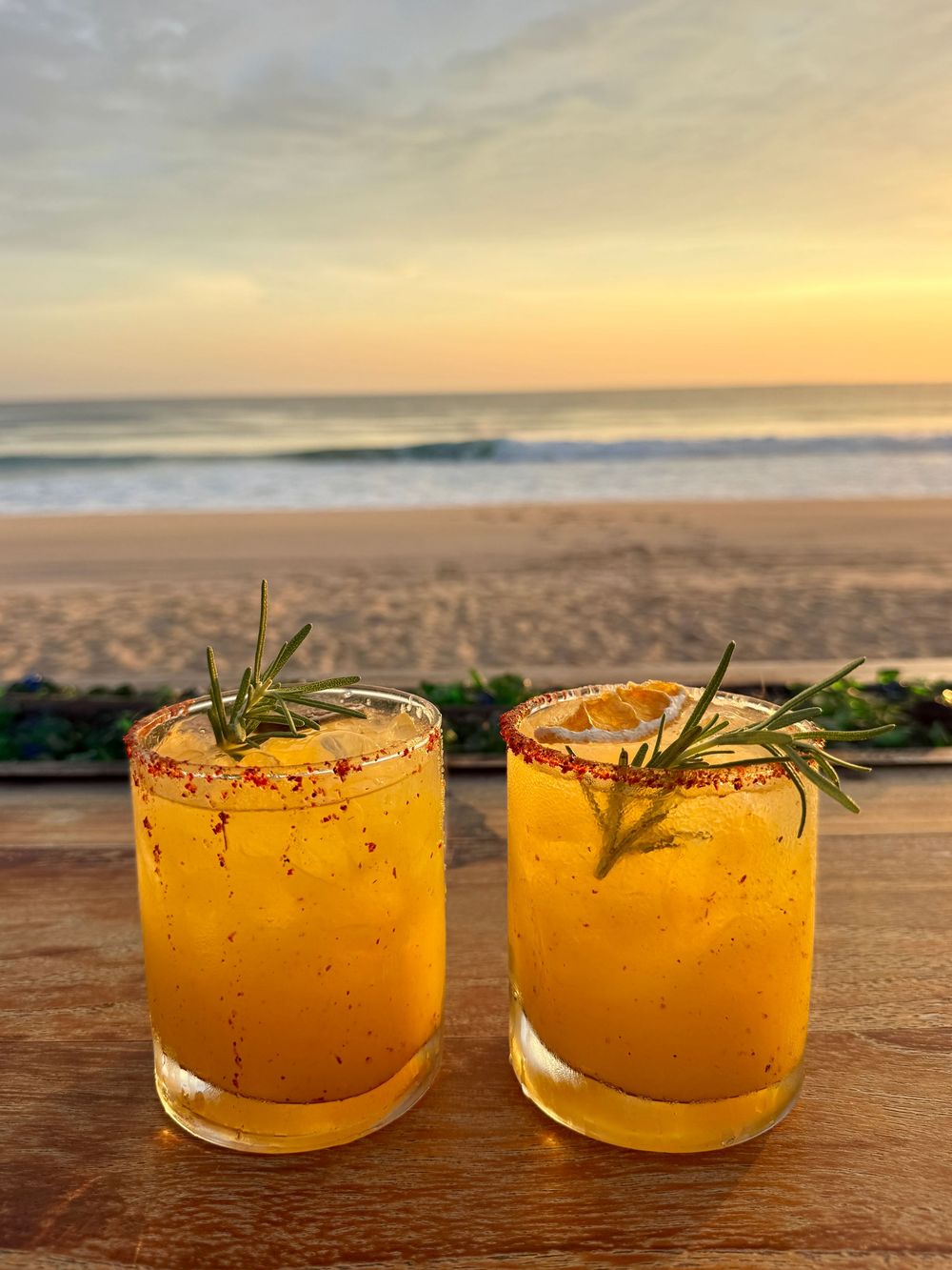 Two spicy mango margaritas with chili-salt rims and rosemary garnish on a wooden bar overlooking a sunset beach.