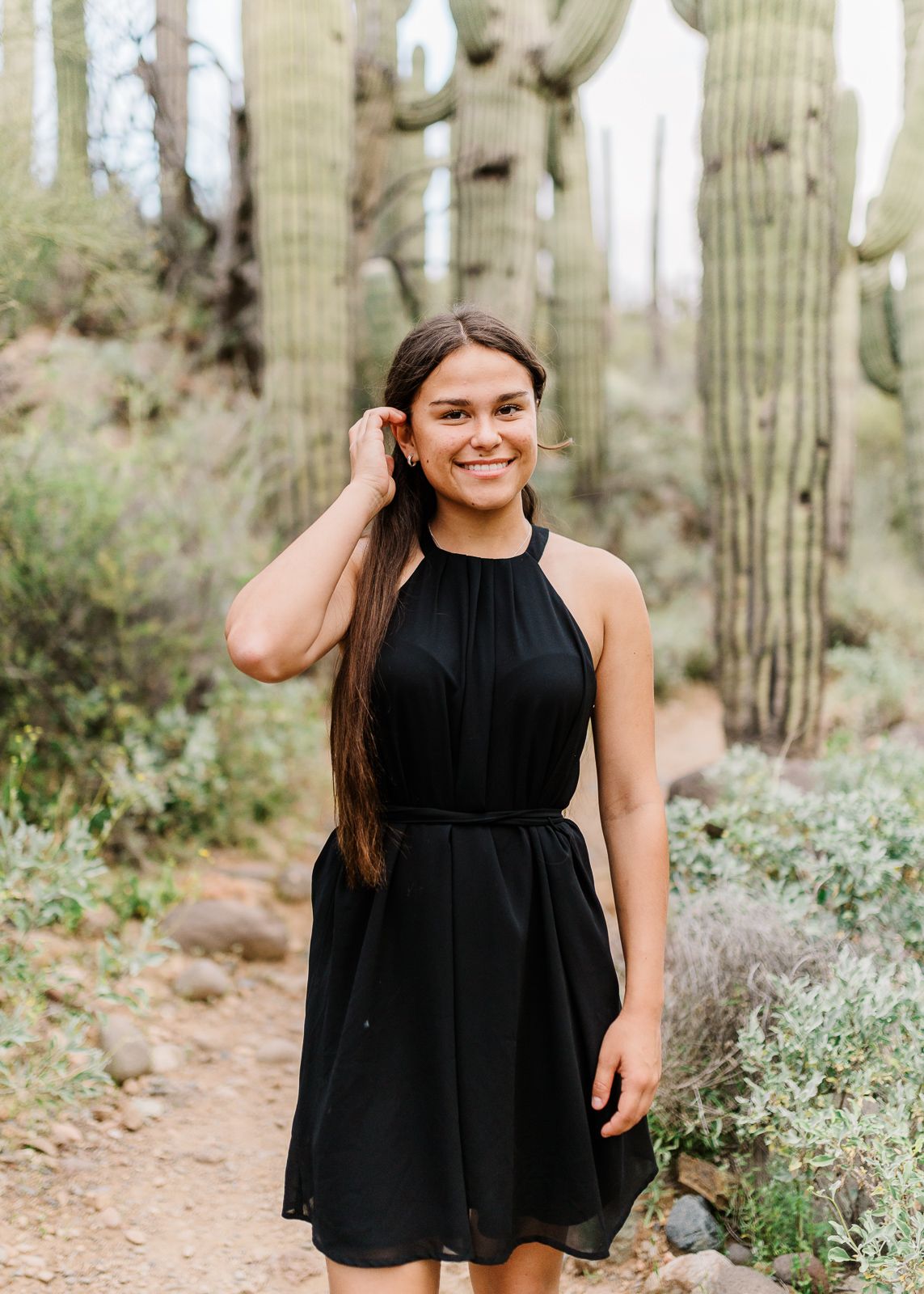 Young woman in black halter dress smiling on a desert trail surrounded by tall saguaro cacti.